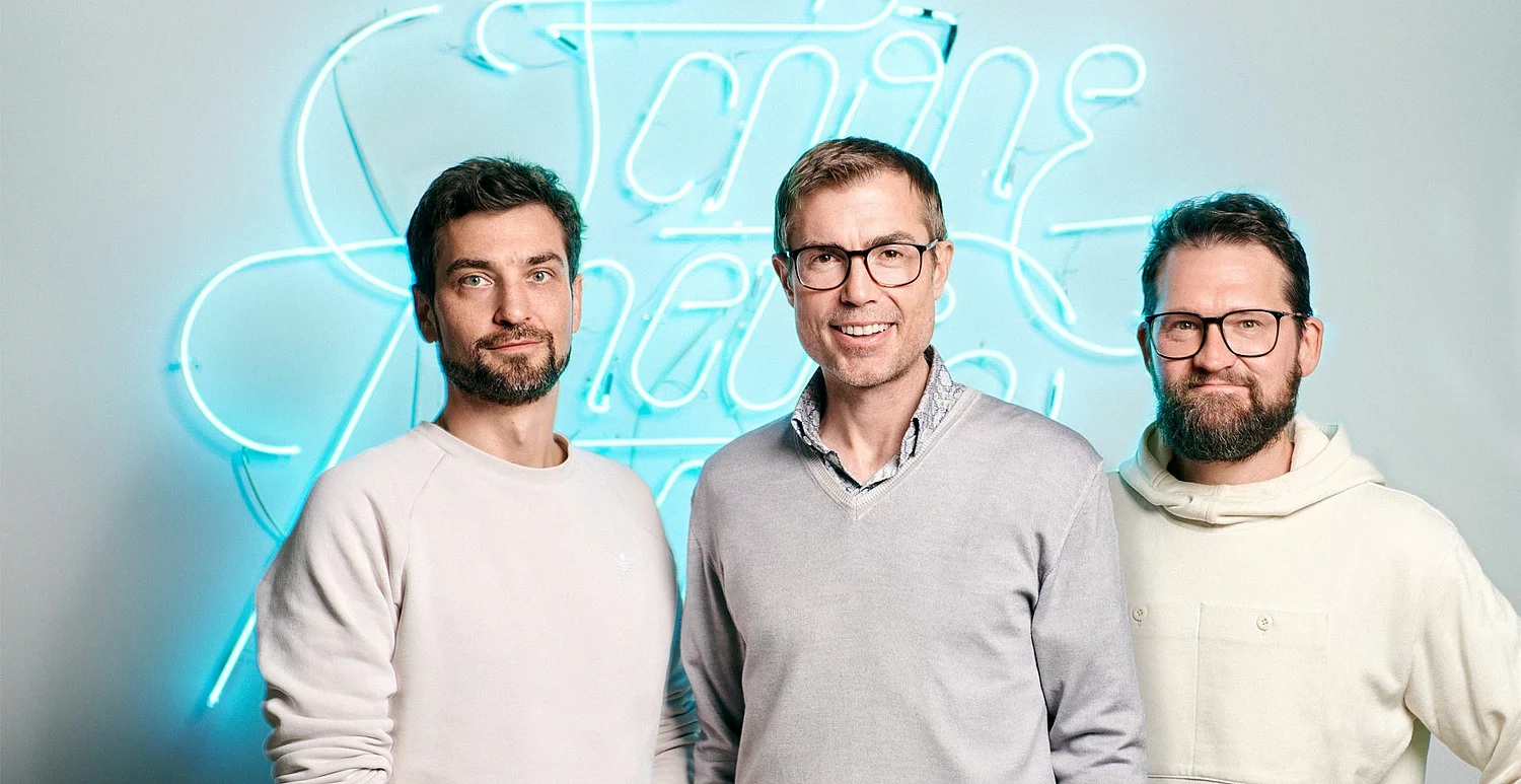 Ein Bild von Drei Männern, die nebeneinander vor einer SNK Neon-Leuchtschrift im SNK-Büro stehen